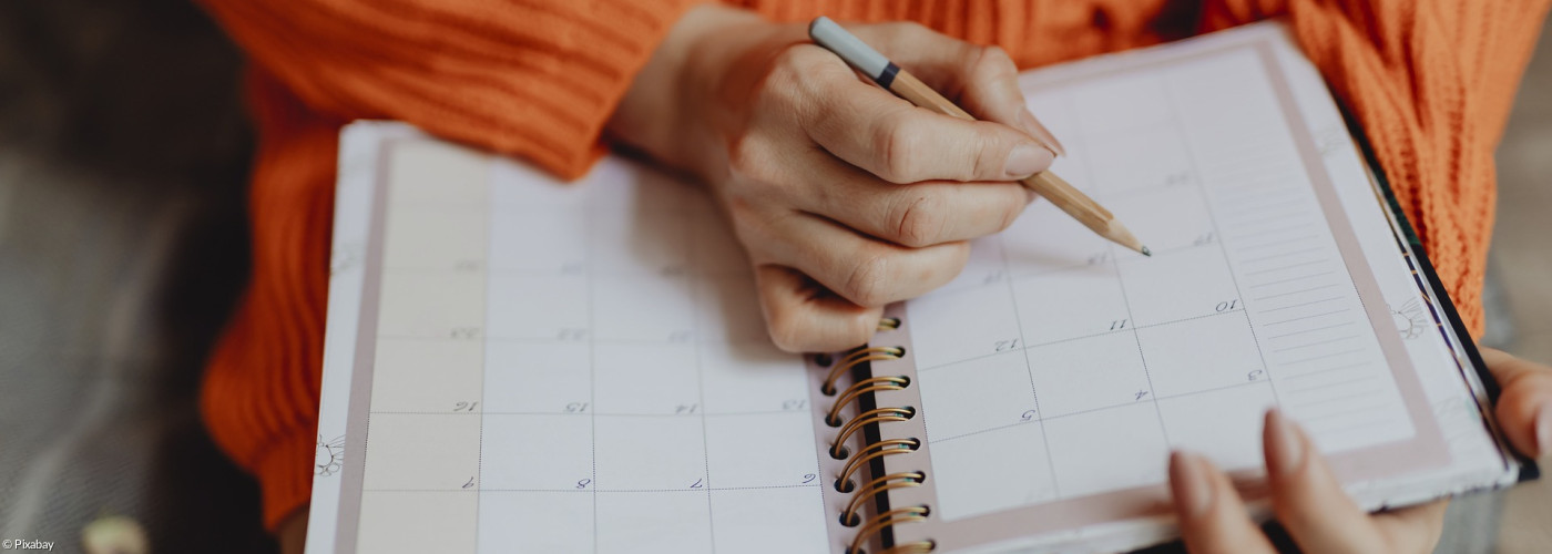 Eine Frau in orangem Pullover notiert sich einen Termin in einem aufgeschlagenen Programmkalender