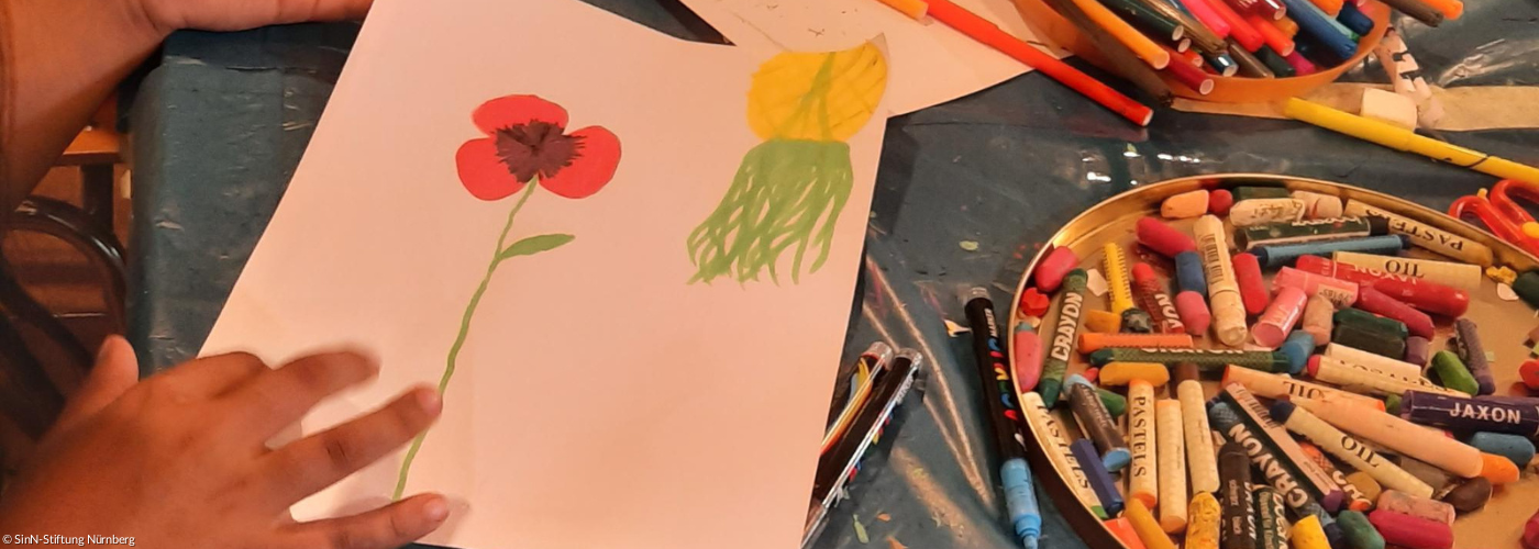 Eine Kinderhand mit einem Buntstift um ein Bild mit einer roten Blume zu zeichnen, daneben ganz viele weiter Buntestifte