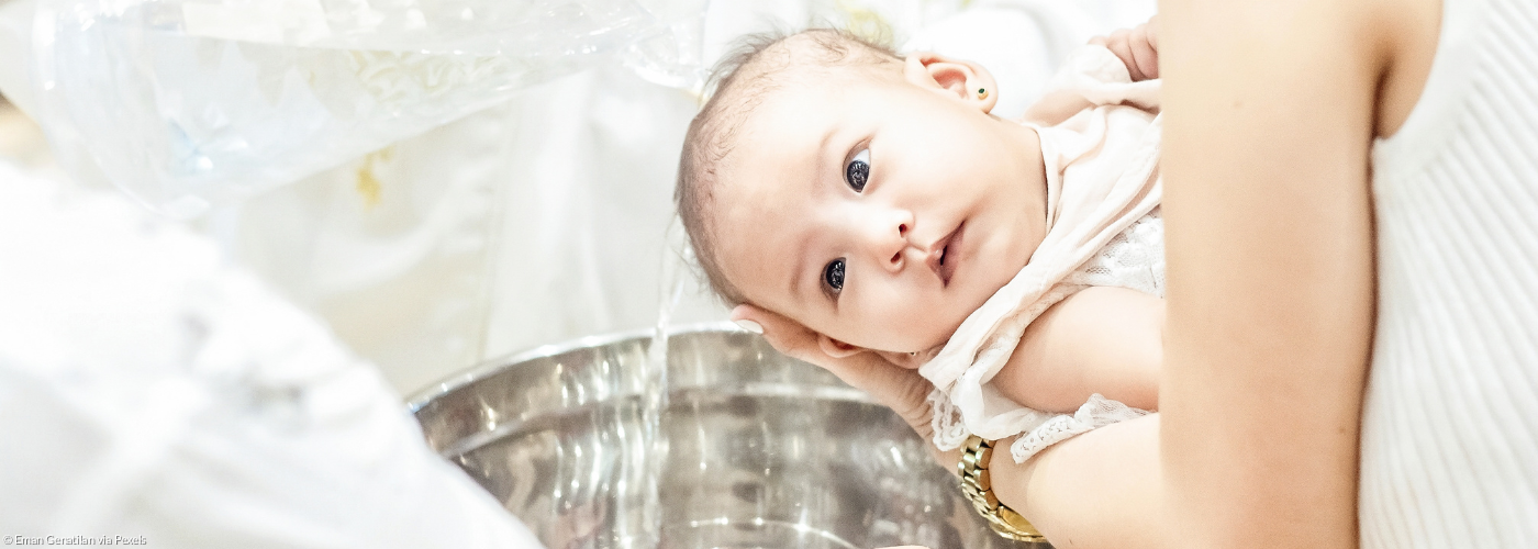 Ein Säugling wird in weißem Taufgewand von seiner Mutter über den Taufstein gehalten