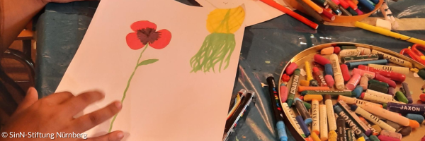 Eine Kinderhand mit einem Buntstift um ein Bild mit einer roten Blume zu zeichnen, daneben ganz viele weiter Buntestifte